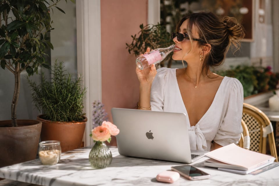 A woman at a sunny cafe sipping Holy Hana Water beside her laptop